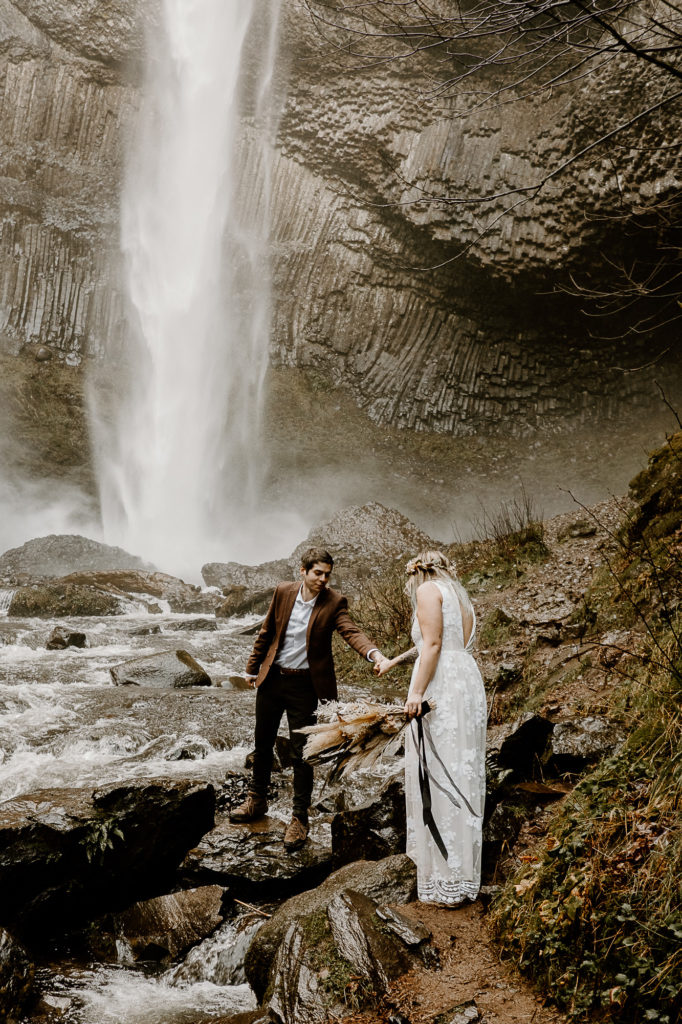 bride and groom walking along rocks by waterfall
