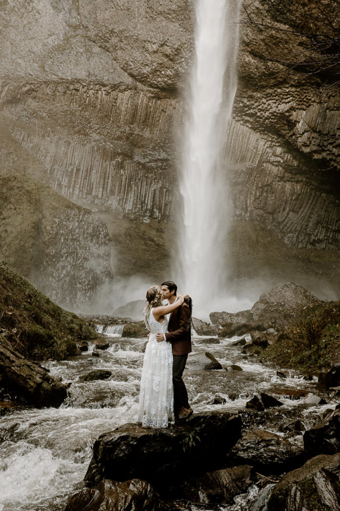 bride and groom kissing by a waterfall on a rock