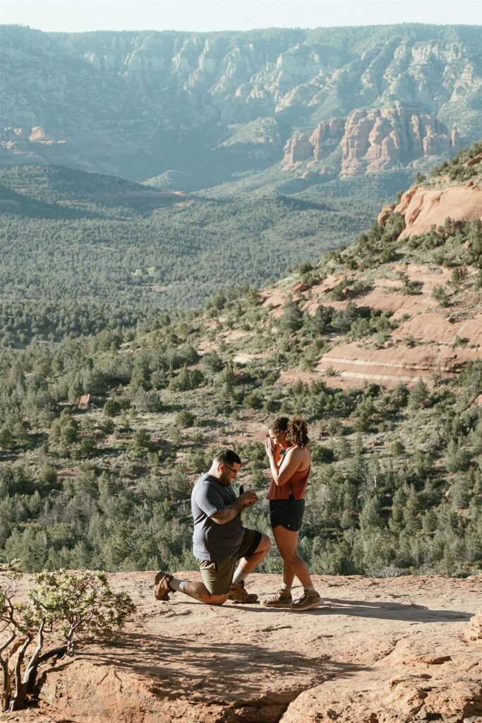 "A heartfelt surprise proposal at Sedona's Devil's Bridge, capturing the emotional moment as he gets down on one knee with stunning desert views in the background."