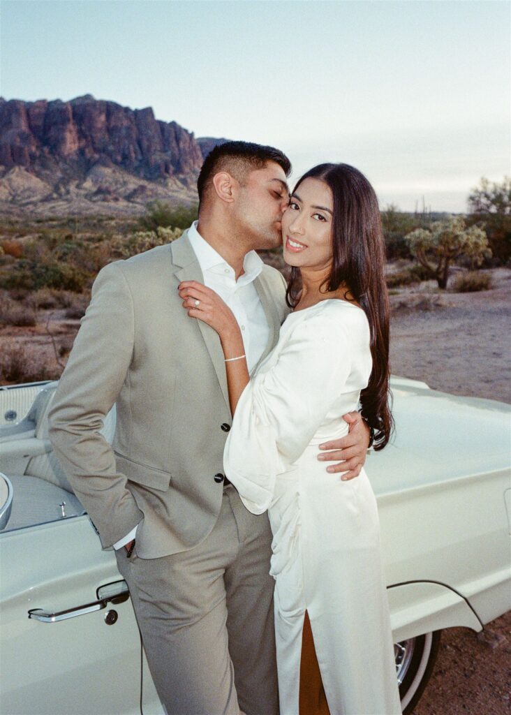 Man kissing woman on the cheek in the desert in front of a vintage car